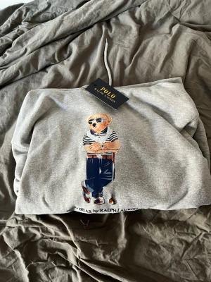 Grå hoodie Polo Ralph Lauren björn - Grå hoodie från Polo Ralph Lauren med ikoniskt björntryck framtill. Klassisk huva med dragsko och mjukt material i bomullsblandning. Perfekt för en chill och stilren look. Snygg detalj med björnen i solglasögon och randig tröja.