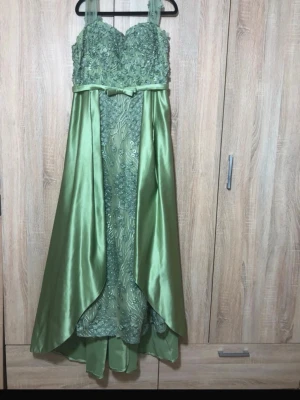 Grön långklänning med spets och satin - Elegant grön långklänning med spetsdetaljer upptill, smala axelband och vacker rosettdetalj i midjan. Kjolen är i glansigt satinmaterial med lager och broderade blommor framtill. Perfekt för bal eller festliga tillfällen. Pris kan diskuteras:) skriv gärna mått så att man kan veta ifall det passar eller inte