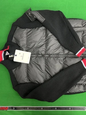 Svart dunväst från Moncler - Snygg svart dunväst från Moncler med stickade ärmar och quiltad front. Dragkedja framtill och ribbade muddar med röd och vit detalj. Klassisk Moncler-logga på ärmen och en modern, sportig look. Perfekt för lager-på-lager och streetstyle.