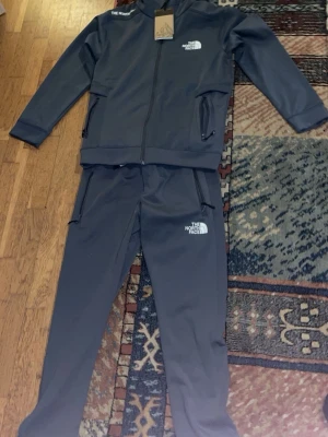 Grått tracksuit från The North Face - Snyggt grått tracksuit-set från The North Face med dragkedja på jackan och fickor med dragkedja både på överdel och byxor. Setet har logga på bröstet och låret samt ribbade muddar. Perfekt för chill dagar hemma eller när du vill vara bekväm./ köpt i Qatar airport för 2100 den e helt ny kan man säga har bara haft på mig den en gång pris kan diskuteras vid snabb affär