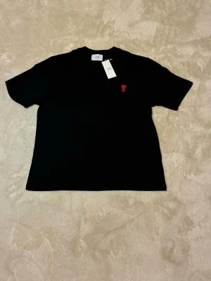 Svart t-shirt från Ami Paris - Snygg svart t-shirt från Ami Paris med klassisk passform och korta ärmar. På bröstet finns Ami Paris ikoniska röda hjärtdetalj. Tillverkad i mjuk bomull som känns skön mot huden. Perfekt för dig som gillar stilrena plagg med en twist.