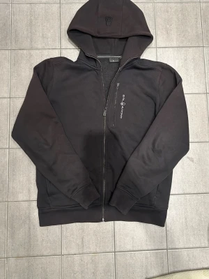 Svart zip-hoodie från Sail Racing - Svart hoodie med dragkedja från Sail Racing. Modellen har huva, broderad logga på bröstet och är tillverkad i en mjuk bomulls- och polyestermix. Perfekt för en avslappnad och sportig stil.