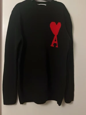 Svart stickad tröja från Ami Paris - Svart stickad tröja från Ami Paris med röd hjärtlogga och bokstaven A på bröstet. Tröjan har rund halsringning och långa ärmar. Perfekt för dig som gillar stilrena plagg med en twist.