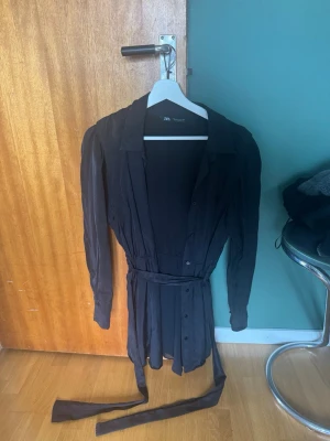 Svart skjortklänning från Zara - Stilren svart skjortklänning från Zara med långa ärmar, knappar framtill och avtagbart midjebälte. Klänningen har klassisk krage och är tillverkad i ett mjukt, lätt tyg som ger en snygg passform. Perfekt för dig som gillar en enkel men trendig look.