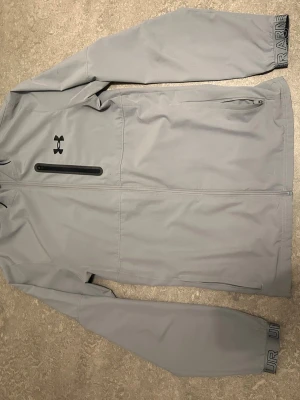 Ljusgrå vindjacka från Under Armour - Snygg ljusgrå vindjacka från Under Armour med dragkedja framtill, svart logga på bröstet och diskreta detaljer på ärmsluten. Jackan har en bröstficka med dragkedja och är tillverkad i ett lätt och smidigt material som passar perfekt för blåsiga dagar.