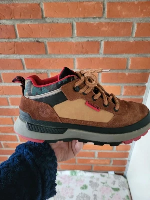 Timberland Field Trekker Skor Brun Strl 42 - Säljer ett par snygga Timberland kängor i brunt mocka och beige textil med svarta och röda detaljer. Skorna har grov sula i grått och rött, samt snörning och camo-mönstrad kant upptill. Perfekta för dig som vill ha en cool och robust look.