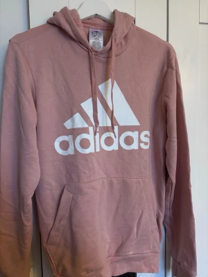 Rosa hoodie från Adidas med logga - Säljer en rosa hoodie från Adidas med stor vit logga på bröstet. Hoodien har huva med dragsko, magficka och ribbade muddar. Tillverkad i mjuk bomullsblandning, perfekt för en avslappnad och sportig stil.
