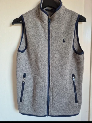 Grå Ralph Lauren Fleeceväst - Snygg grå väst från Polo Ralph Lauren i ullmix med marinblå detaljer. Västen har hög krage, två fickor med dragkedja och stängs med dragkedja framtill. Klassisk broderad logga på bröstet och marinblå kantband längs ärmhål och nederkant. Säljer nu denna ruggigt feta väst då den va för liten, den är i ett fint skick, storlek S och passar dig mellan 170-180 Max. Hör av er vid frågor/liknande✌🏻