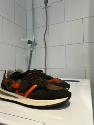 PM (Philip model) sneakers med orange detaljer - Säljer ett par sneakers med kamouflagemönster i grönt, brunt och orange. Skorna har svarta mockadetaljer, vita sulor och svarta snören. Perfekta för dig som vill sticka ut med en unik stil. Materialmix av mocka och textil. Pris kan sänkas vid snabb affär skickas samma dag om det är under måndag till fredag 