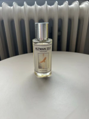 Altman 22.0 Eau de Parfum - En stilren parfymflaska i glas med metallock från One Bold Obsession. Doften heter Altman 22.0 och flaskan har en minimalistisk etikett med en fågel i guldton. Innehållet är ljusgult och flaskan har en modern, cylindrisk form.