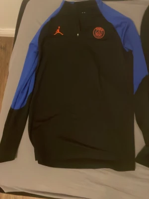 PSG x Jordan svart träningsset - Säljer ett svart PSG x Jordan träningsset med långärmad tröja och byxor. Tröjan har blå ärmar, halv dragkedja och röda loggor på bröstet. Byxorna har röda PSG- och Jordan-loggor samt Paris-tryck längs benet. Perfekt för fotbollsträning eller chill.