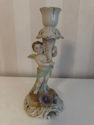 Ljusstake porslin med ängel och blomma - Dekorativ ljusstake i porslin med en ängel som håller i ljushållaren. Basen är utsmyckad med en lila blomma och gulddetaljer. Färgerna går i vitt, grönt, lila och guld. Perfekt som prydnad eller till middagsbordet.