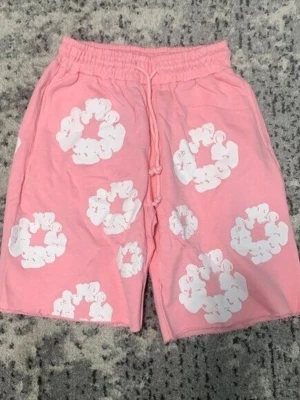Rosa shorts med vita blommor - Säljer ett par rosa shorts med vita blommönster och resår i midjan samt snörning framtill. Shortsen har en avslappnad passform och är perfekta för varma dagar. Materialet känns mjukt och bekvämt, troligen bomull eller bomullsblandning.
