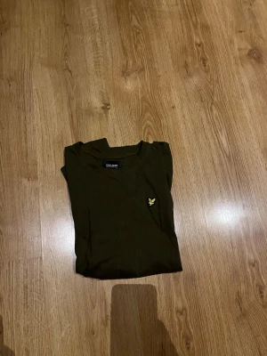 Olivgrön t-shirt från Lyle & Scott - Snygg olivgrön t-shirt från Lyle & Scott med klassisk rund hals och ikonisk gul logga broderad på bröstet. Tillverkad i mjuk bomull för en bekväm känsla. Perfekt basplagg för en avslappnad stil.