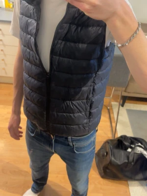 Mörkblå dunväst från Uniqlo - Säljer en mörkblå dunväst från Uniqlo, modell Ultra Light Down. Västen är lätt och smidig med quiltad design och dragkedja framtill. Perfekt att slänga över en hoodie eller t-shirt för extra värme. Tillverkad i Bangladesh.