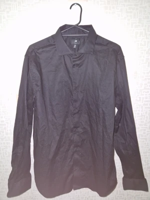Svart slim fit skjorta från H&M - Klassisk svart skjorta från H&M i slim fit-modell. Skjortan har långa ärmar, knappar framtill och en stilren krage. Perfekt för dig som gillar en enkel och clean look. Tillverkad i ett mjukt bomullstyg som känns bekvämt hela dagen. Bara använd 1 gång 
