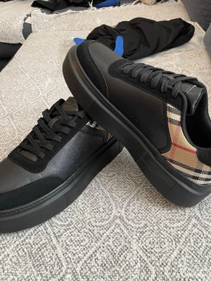 Burberry sneakers med rutigt mönster - Snygga sneakers från Burberry med klassiskt beige, svart och rött rutmönster på sidorna. Ovandelen är i svart läder och textil, med svarta skosnören och robust svart sula. Perfekta för dig som vill ha en stilren och trendig look.