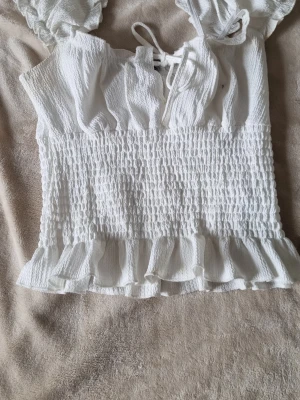 Vit volangtop från Gina Tricot - Supersöt vit topp från Gina Tricot med volangkant nedtill, smockad midja och knytning fram. Några små fläckar som kommer förmodligen försvinna i tvätten bild på skickas vid intresse