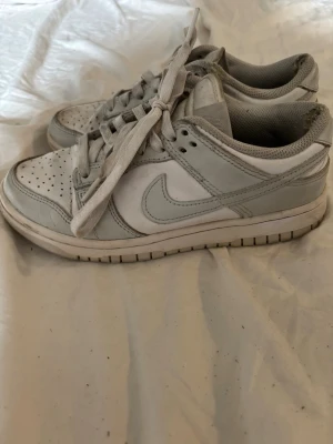 Nike Dunk Low vita/grå sneakers - Säljer ett par Nike Dunk Low sneakers i vitt och ljusgrått läder. Klassisk siluett med perforerad tå låda medföljer ej, vita skosnören och den ikoniska Swoosh-loggan på sidan. Perfekta för dig som gillar stilrena och tidlösa sneakers.