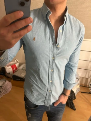 Ljusblå skjorta från Ralph Lauren - Säljer en ljusblå skjorta från Ralph Lauren med klassisk button-down krage och broderad logga på bröstet. Skjortan har långa ärmar, vita knappar och är tillverkad i ett mjukt bomullstyg. Perfekt för en clean och stilren look.