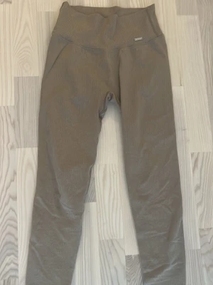 Ribbade bruna leggings från Aim'n - Säljer ett par ribbade leggings från Aim'n i en snygg brun färg. De har hög midja och är tillverkade i ett stretchigt material som sitter skönt på kroppen. Perfekta för både träning och chill. Metallogo framtill vid linningen för extra detalj.