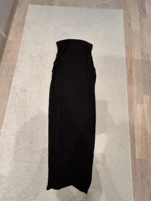 Svart långkjol med rynkade sidor - Säljer en svart långkjol med rynkade detaljer längs sidorna. Kjolen har en rak och smal passform som ger en clean och stilren look. Perfekt att styla med en crop top eller en oversized tröja. Materialet känns mjukt och stretchigt.