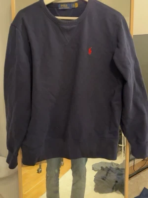 Mörkblå stickad tröja Polo Ralph Lauren - Mörkblå stickad tröja från Polo Ralph Lauren med rund halsringning och röd broderad logga på bröstet. Tröjan har långa ärmar och ribbade muddar vid ärmslut och nederkant. Perfekt för en avslappnad och stilren look.