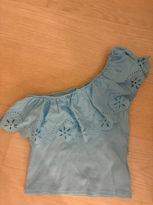 Ljusblå offshoulder topp med volang - Trendig ljusblå offshoulder topp med bred volangkant och broderade blommor. Toppen är ribbad och har en axelbandslös design som ger en snygg och somrig look. Perfekt för dig som vill sticka ut med en söt och unik topp. Storleken 13 - 14 år ungefär en xs