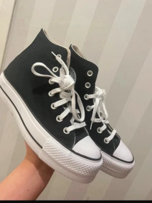 Svarta Converse Chuck Taylor High - Klassiska svarta Converse Chuck Taylor All Star high sneakers med vit tåhätta, vita snören och sula. Tillverkade i canvas med gummisula och metallöljetter. Ikonisk logga på sidan och tidlös design som alltid funkar.