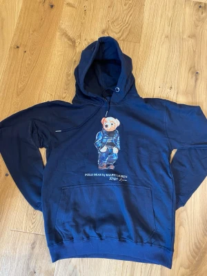 Marinblå hoodie Polo Bear Ralph Lauren - Säljer en marinblå hoodie från Ralph Lauren med den ikoniska Polo Bear-trycket på bröstet. Hoodien har huva med dragsko, stor magficka och ribbade muddar. Materialet är mjukt och skönt, perfekt för en avslappnad stil.