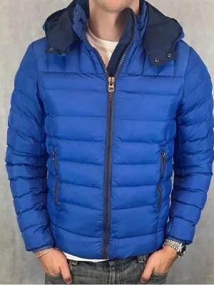 Blå pufferjacka från Colmar - Snygg blå pufferjacka från Colmar med avtagbar huva och två dragkedjeförsedda fickor framtill. Jackan har ett quiltat mönster och Colmar-logga på ärmen. Perfekt för kalla dagar och riktigt skön att bära tack vare det lätta materialet.