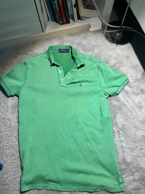 Grön pikétröja från Polo Ralph Lauren - Snygg grön pikétröja från Polo Ralph Lauren med klassisk krage och knappar framtill. Tröjan har det ikoniska broderade logotypen på bröstet och är tillverkad i mjuk bomull. Perfekt för en avslappnad men stilren look.