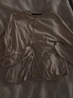 Beige kofta från 157 - Ljusbeige kofta från 157 med rund halsringning och knäppning framtill. Tillverkad i ett mjukt, tunt material med ribbade muddar vid ärmslut och nederkant. Enkel och stilren design som passar till många olika outfits. Fint skick! Använd 2 gånger.