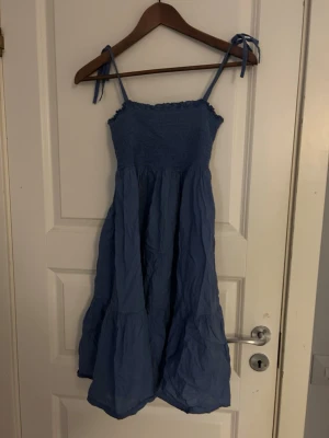 Blå kort klänning - Säljer en blå klänning från Ellos i storlek 34/36. Klänningen har knytband vid axlarna och smock upptill, vilket ger en skön och avslappnad passform. Materialet är mjukt och lätt, perfekt för varmare dagar. Den har en volangig och rynkad nederdel som ger extra rörelse.