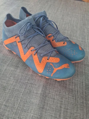 Blå-orange fotbollsskor från Puma - Snygga fotbollsskor från Puma i blått med orange detaljer och logga. Skorna har stickad strumpliknande krage, snörning och greppvänlig sula med dobbar för spel på gräs. Perfekta för dig som vill sticka ut på planen.