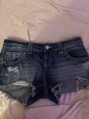 Mörkblå slitna jeansshorts - Säljer ett par mörkblå jeansshorts med slitna detaljer och fransig kant. Shortsen har klassiska fem fickor, vita kontrastsömmar och dekorativa nitar på bakfickorna. Perfekta för sommaren och ger en cool, avslappnad vibe.