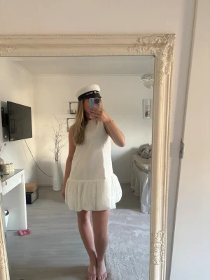 Vit kort klänning med volang - Säljer en vit kort klänning med volang nedtill. Klänningen är ärmlös och har en enkel, rak passform. Materialet ser ut att vara bomull eller polyester med en lätt struktur. Perfekt för student eller sommarens alla tillfällen. Har som paljetter i sig 