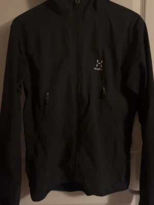 Svart vindjacka från Haglöfs - Svart vindjacka från Haglöfs med huva och hel dragkedja framtill. Jackan har två snedställda fickor med dragkedja och diskret logga på bröstet. Tillverkad i ett lätt och smidigt material som skyddar mot vind. Perfekt för utomhusaktiviteter. Vädigt fint skick, inga skador eller liknande. Storlek M men passar också S.