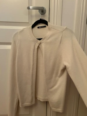 Beige kofta från Gina Tricot - En stilren krämvit kofta från Gina Tricot med rund halsringning och ribbade kanter. Koftan har en enkel knäppning upptill och är tillverkad i ett mjukt, stickat material. Perfekt att slänga över en t-shirt eller klänning.