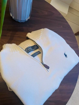 Vit halvzip-tröja från Polo Ralph Lauren - Snygg vit stickad tröja från Polo Ralph Lauren med halv dragkedja och broderad logga på bröstet. Tröjan är i bomull och har ribbade muddar vid krage och ärmslut. Perfekt för en stilren och klassisk look. Cremevit färg 