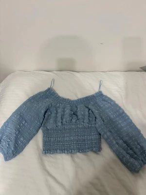 Blå offshoulder topp med puffärmar - Trendig ljusblå topp med offshoulder-design och långa puffärmar. Smockad midja och strukturmönster i tyget ger en cool och unik look. Tunt axelband för extra detalj. Perfekt för dig som gillar en söt och modern stil.