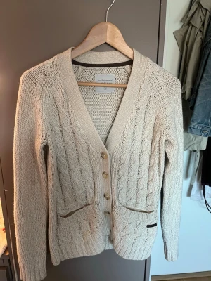 Beige kabelstickad kofta med mockadetaljer - Beige kabelstickad kofta från peakPerformence, bruna knappar och två fickor framtill. Mjukt material och ribbade muddar. Nästan ny skick och super fin, men passar inte i färgen 