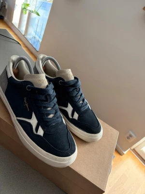Mörkblåa sneakers från Pepe Jeans - Snygga mörkblå sneakers från Pepe Jeans med vit sula och vita detaljer på sidan. Ovandelen är i mocka och textil, med grå hälparti och beige plös. Klassisk låg modell med snörning.                                       Original box kommer med!