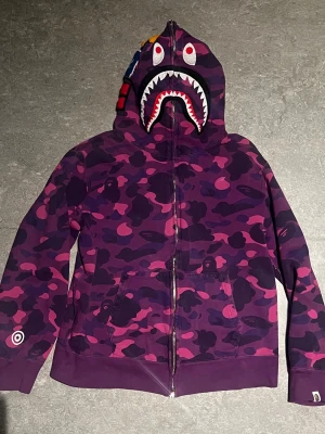 Purple BAPE Zip-Up hoodie - Säljer en ikonisk BAPE hoodie i lila färg! Dragkedjan går hela vägen över huvan.  broderade bokstäver, tiger detalj och hajdetaljer på huvan! storlek XL men sitter som large! Kommer med dustbag