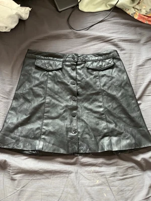 Svart skinnkjol från H&M med knappar - Snygg svart kort skinnkjol från H&M med knappar framtill och två dekorativa ficklock vid midjan. Kjolen har en lätt A-linjeform och är perfekt för att skapa en edgy look. Materialet är syntetiskt skinn som ger en cool vibe.