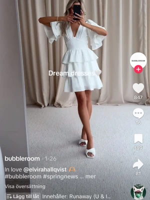 Vit volangklänning från Bubbleroom - Säljer en vit kortklänning från Bubbleroom med v-ringad omlottfront, volanger och vida kortärmar. Klänningen har en feminin och luftig design som passar perfekt till ljusa säsonger.