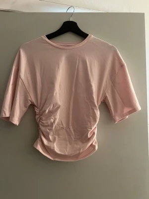 Ljusrosa topp med rynkdetalj - Trendig ljusrosa topp med korta ärmar och snygg rynkdetalj på ena sidan. Tillverkad i mjuk bomull som känns skön mot huden. Perfekt för dig som gillar en enkel men ändå unik stil.