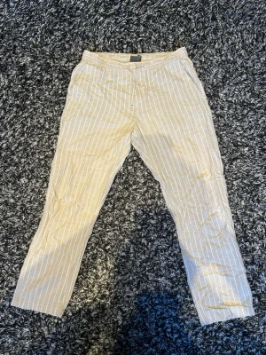 Beige linnebyxor med vita ränder - Snygga beige linnebyxor med tunna vita vertikala ränder och dragsko i midjan. Byxorna har raka ben och sidofickor, perfekta för en avslappnad och trendig stil. Luftigt material som passar bra till varmare dagar.