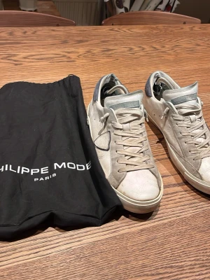 Philippe Model vita sneakers - Snygga vita sneakers från Philippe Model med grå och blå detaljer. Skorna har lågt skaft, klassisk snörning och är tillverkade i läder med mockadetaljer. Ikonisk sköld-logga på sidan och bekväm sula. Perfekta för en trendig streetstyle-look.