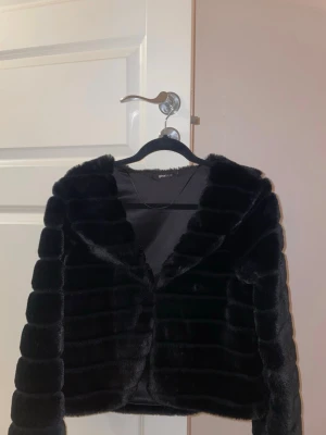 Svart pälsjacka från Gina Tricot - Snygg svart pälsjacka från Gina Tricot i storlek S. Jackan har en kort, boxig passform med breda, horisontella paneler och stor krage. Perfekt för att ge din outfit en lyxig touch under kalla dagar.